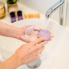 Hände schäumen ein rundes, violettes Stück INUANA Seife unter fließendem Wasser zur sanften Reinigung auf.