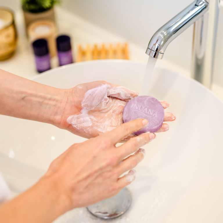 Hände schäumen ein rundes, violettes Stück INUANA Seife unter fließendem Wasser zur sanften Reinigung auf.