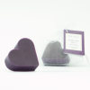 Eine herzförmige violette Seife von INUANA neben einem verpackten Duftseife-Herz im Organzasäckchen.