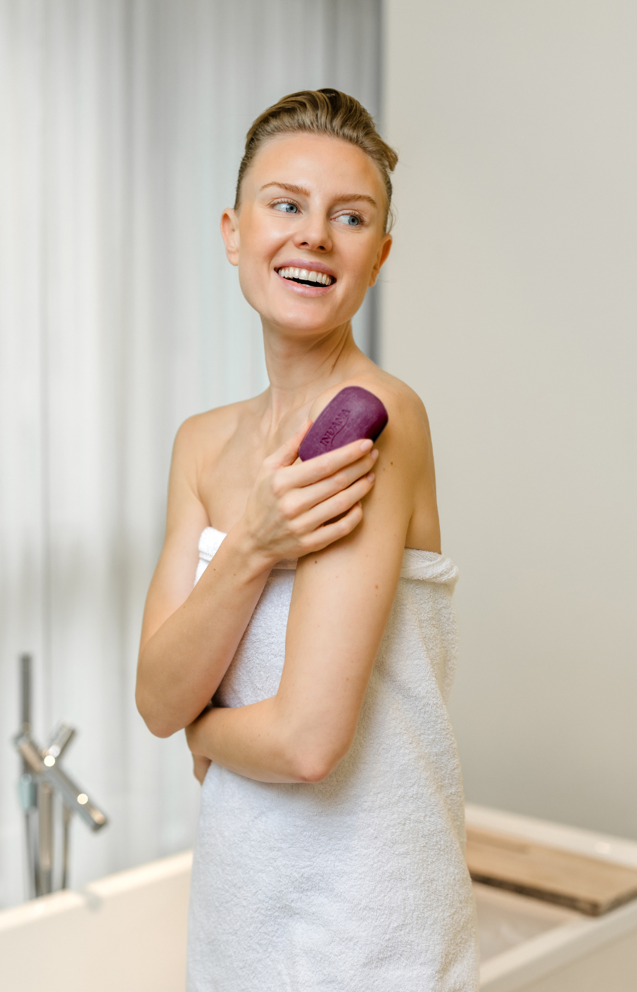Eine lächelnde Frau im weißen Handtuch hält das violette INUANA Duschpflege-Stück sanft an ihre Schulter.