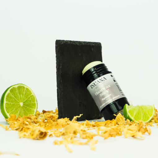 Geöffneter Haarwax Stick Signature von INUANA vor Schiefertafel mit Limetten und Holzspänen