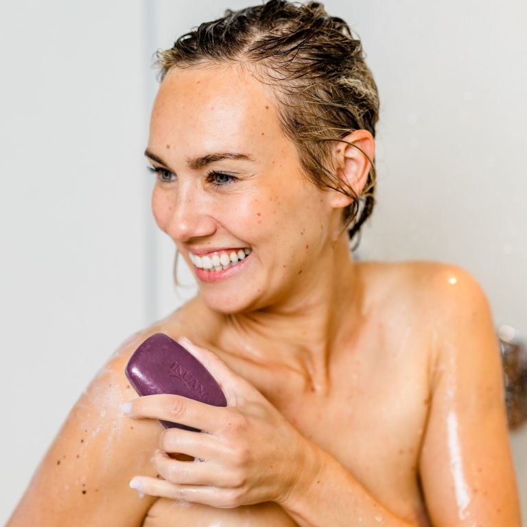 Eine lächelnde Frau unter der Dusche nutzt das lila INUANA Duschpflege-Stück für sanfte Körperpflege.