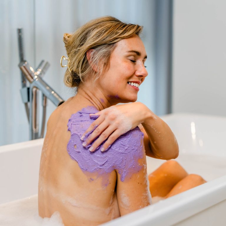 Eine Frau trägt die lila INUANA Shower Mousse Duschpflege als sanftes Peeling auf ihre Schulter auf.