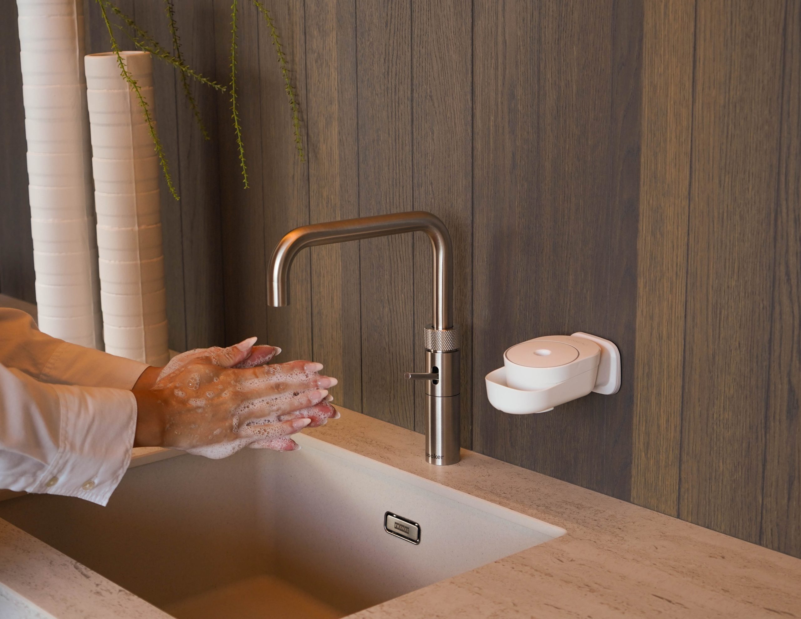 Eingeseifte Hände am Waschbecken neben einem weißen INUANA Wanddispenser für pflegende Seife.