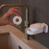 Hand hält runde feste Seife vor weißem INUANA Wanddispenser am Waschbecken für nachhaltige Händereinigung.