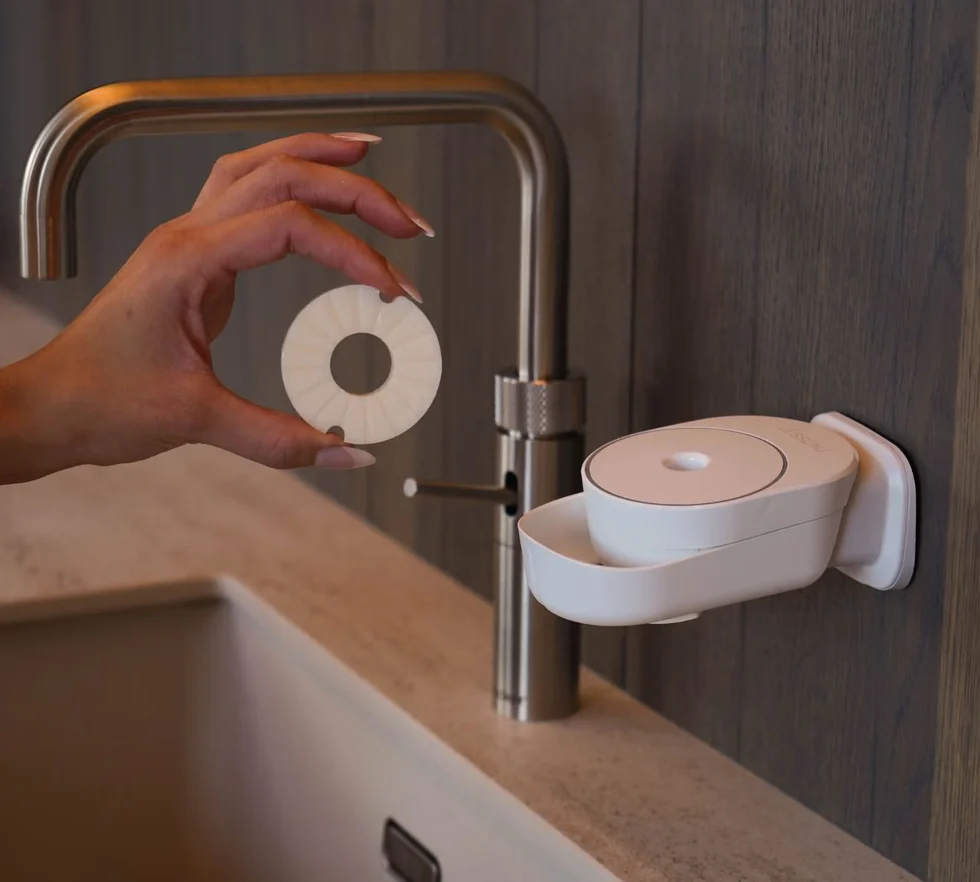 Hand hält runde feste Seife vor weißem INUANA Wanddispenser am Waschbecken für nachhaltige Händereinigung.