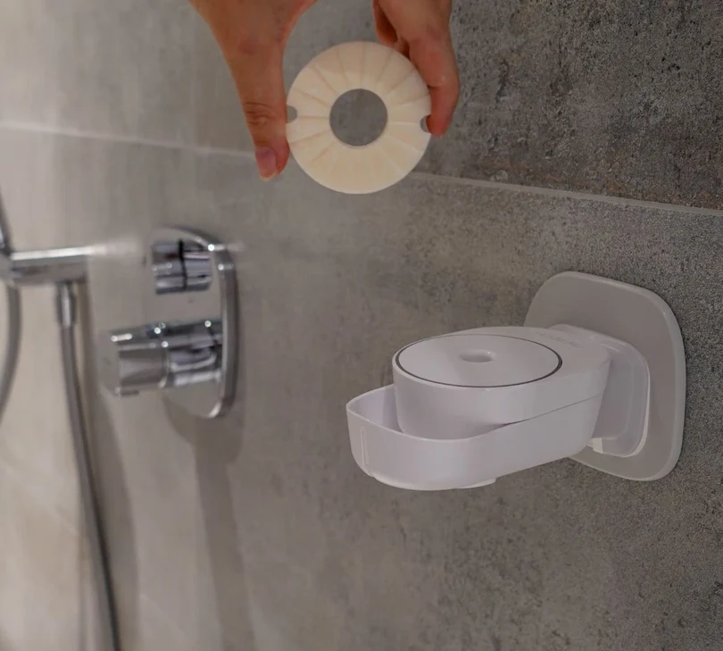Hand hält runde feste Seife über weißem INUANA Wanddispenser an Fliesenwand für Duschpflege.